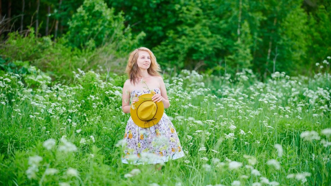 Caucasian woman in a floral dress holding a hat in a green, blooming field on a bright summer day.