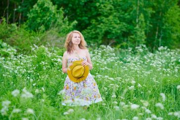 Caucasian woman in a floral dress holding a hat in a green, blooming field on a bright summer day.
